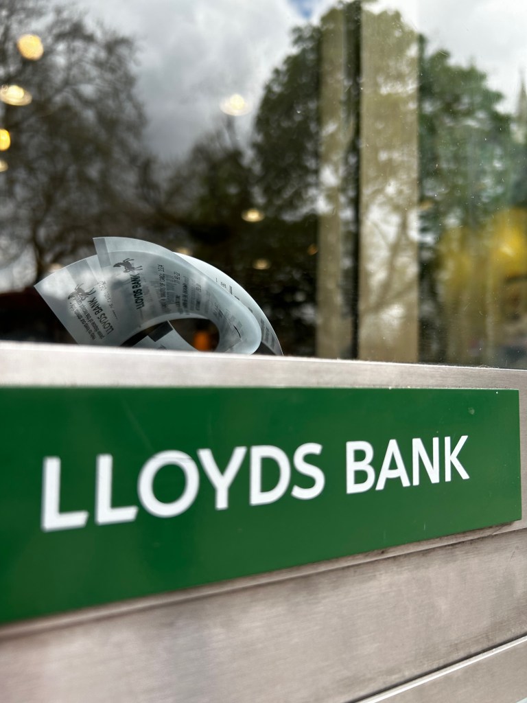 Letterbox from a Lloyds Bank branch.  There are a couple of transaction receipts on top of the letterbox. It helps the reader to understand that migrants need to get a bank account before getting a national insurance number. 