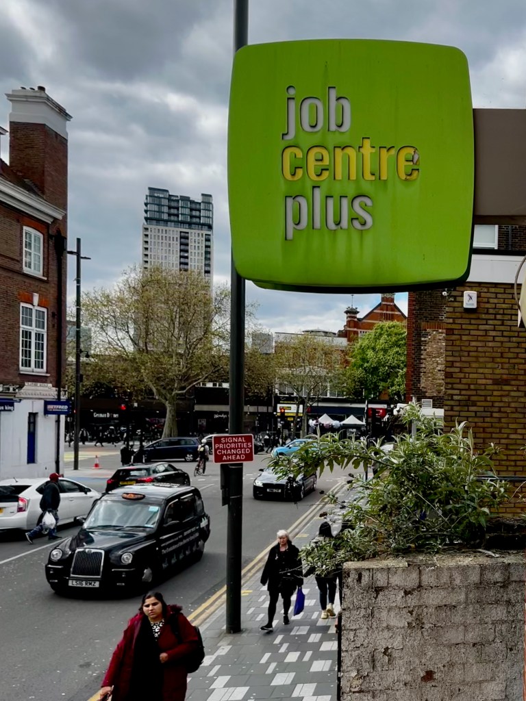 Job Centre Plus sign with people walking on the street as a background. The picture helps the reader to understand that migrants need to go to a job centre plus to get a national insurance number or NINO.